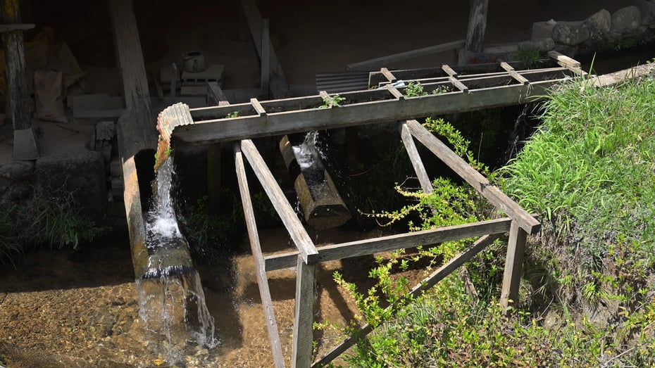 Outside the hut, water from the stream fills a hollowed wooden bucket at the end of the lever. When the bucket becomes heavy, it lifts the pestle; once the water spills, the pestle comes crashing down.